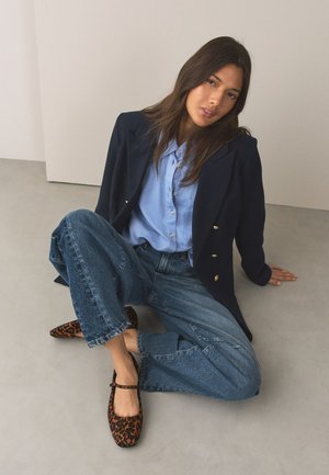 Femme assise par terre portant un blazer bleu marine, une chemise bleu clair à boutons, un jean à jambes larges et des chaussures Mary Jane à motif léopard.