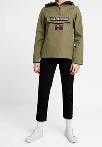 Oliven grønne pulloverjakke med lynlås hals, en frontlomme og et norsk flag lap. Sammen med sorte bukser og hvide sneakers.