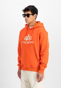 Sudadera con capucha naranja que presenta un bolsillo frontal y un logo blanco. El modelo lleva gafas de sol negras y pantalones cargo claros, mostrando un look casual.