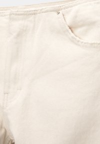 Gros plan sur un tissu en denim écru montrant une poche avant, une passants de ceinture et des détails de rivets en métal sur un jean.