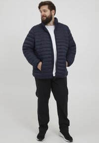 Mornarska puffer jakna s vertikalnim kvadratnim dizajnom, visokim ovratom, zatvaranjem na zip i bočnim džepovima, u paru s crnim hlačama i tenisicama.