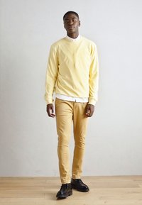 Sweatshirt jaune avec des poignets côtelés, superposé sur une chemise blanche à col. Un pantalon beige et des chaussures noires complètent l'ensemble. Design simple.