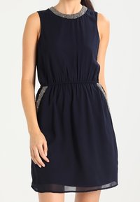 Námořnická modrá bezrukávová šaty s elastickým pasem, s krčkem se stříbrnými akcenty a bočními kapsami, vyrobené z lehkého materiálu s hladkým povrchem.