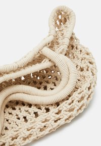 Sac fourre-tout en filet crochet beige avec des poignées tressées épaisses, présentant un motif tricoté ouvert et des bords arrondis sur fond blanc.