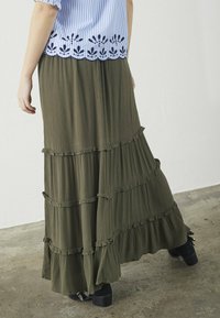 Lang olivengrøn nederdel lavet af krøllet stof, med lagdelte flæser og et svævende design. Bæres med mørke ankelstøvler.