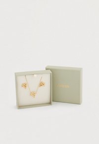 Σετ χρυσών κολιέ με μενταγιόν σε σχήμα καρδιάς, εκτεθειμένα σε ανοιχτό πράσινο κουτί με χρυσή επιγραφή "GUESS". Επένδυση από απαλό ύφασμα.