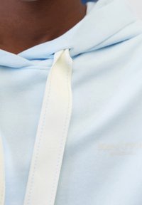 Primo piano di una persona che indossa un hoodie azzurro chiaro con ampie corde bianche e un marchio sobrio sul petto.