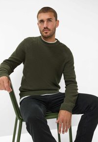 Maglione verde scuro con lavorazione a maglia, scollo rotondo e polsini a costine. Indossato sopra una camicia bianca, abbinato a jeans neri mentre è seduto su una sedia verde.