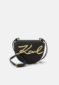 Kruhová černá tkaná taška se zlatým kurzivním logem "Karl", vybavená štíhlým nastavitelným popruhem a texturovaným povrchem.