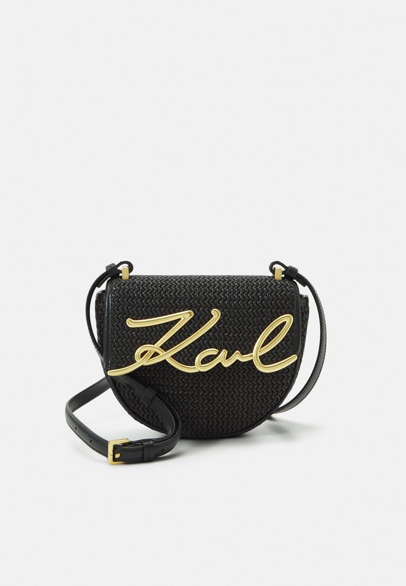 Kruhová černá tkaná taška se zlatým kurzivním logem "Karl", vybavená štíhlým nastavitelným popruhem a texturovaným povrchem.