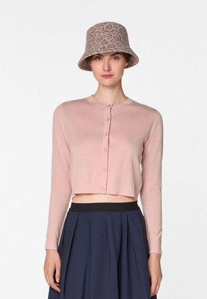 Femme portant un cardigan court à boutons rose clair, une jupe plissée marine, et un chapeau bob beige à imprimé léopard, debout devant un fond blanc.