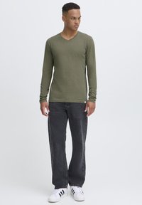 Olivengrünes Langarmshirt mit V-Ausschnitt aus leichtem Stoff, kombiniert mit dunklen Jeans und weißen Sneakers mit schwarzen Streifen.
