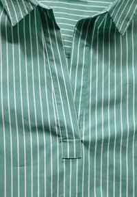 Bluzka w paski z kołnierzykiem w jasnym zielonym kolorze z białymi pionowymi paskami. Miękka tekstura, dekolt w kształcie litery V oraz detal w postaci patki poniżej kołnierza.