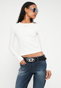 Biele tričko s dlhým rukávom má na hrudi malé logo. Modelka nosí tmavomodré riflové nohavice s čiernym opaskom a striebornými kovovými detailmi. Priehľadné slnečné okuliare.