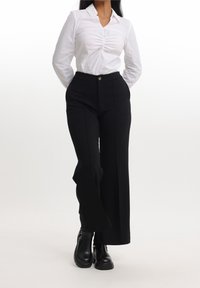Camicia bianca a maniche lunghe con dettaglio plissettato sul davanti, abbinata a pantaloni neri a vita alta e gamba larga e stivaletti neri.