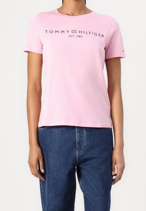 Person trägt ein hellrosa kurzärmliges T-Shirt mit dem Schriftzug "TOMMY HILFIGER EST. 1985" und blaue hoch taillierte Jeans.