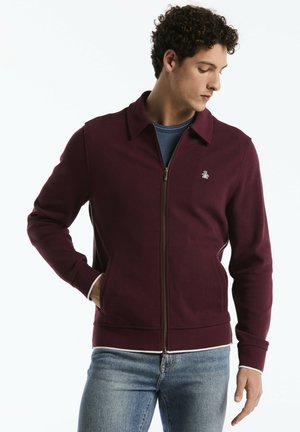 Man wearing a zipped maroon jacket with a small logo on the chest, blue shirt underneath, and light blue jeans, looking down to the side.