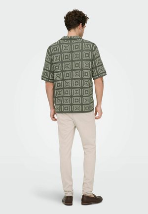 Bărbat stând cu fața spre spate, purtând o cămașă cu mânecă scurtă verde cu model, pantaloni bej și mocasini maro pe un fundal simplu.