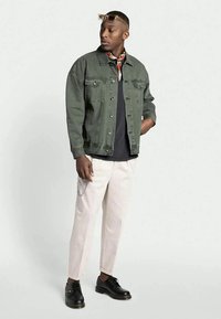 Casaco de ganga verde-oliva com fechos de botão, tshirt preta, calças creme e sapatos pretos de atacador. Inclui um lenço de pescoço estampado e detalhe de corrente.