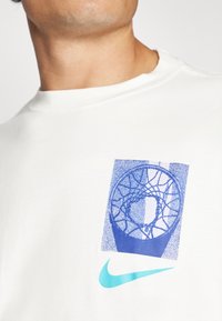 White cotton t-shirt featuring a blue graphic of a basketball hoop and a turquoise Nike logo on the lower front. Soft texture, round neck.