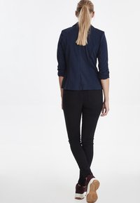 Mörkblåt åtsittande kavaj med uppvikta ärmar, kombinerad med svarta skinny jeans och burgundy sneakers, som visar en avslappnad, lager-på-lager stil.