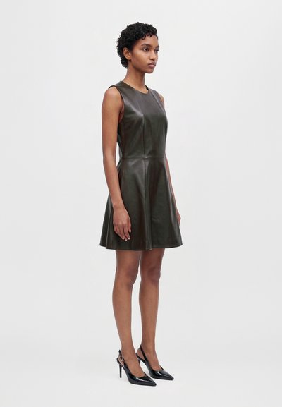 Une femme porte une robe patineuse sombre sans manches, d’aspect cuir, et des escarpins noirs à bout pointu, sur un fond clair uni.