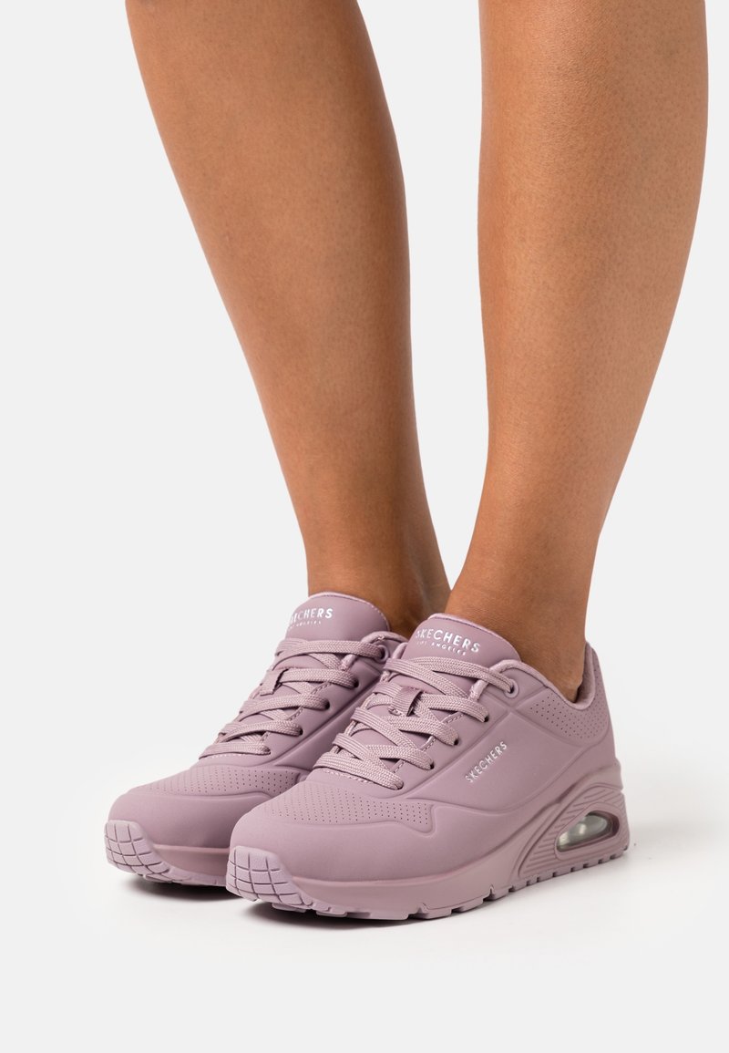 Pieds portant des baskets Skechers violet pastel avec des semelles à coussin d'air visibles, sur un fond clair uni.