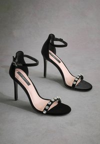 Sandali con tacco alto in suede nero con cinturini alla caviglia, caratterizzati da una parte superiore nera decorata con strass e suole interne rosa.