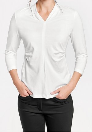 Žena v bílém dlouhém rukávu s výstřihem do V a černých kalhotách s rukama zasunutýma v předních kapsách, stojící před jednobarevným pozadím.