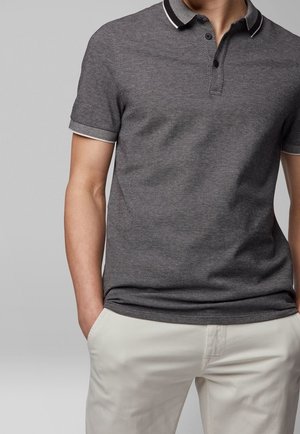 Hombre vestido con una camisa tipo polo de manga corta de color gris oscuro con ribete blanco en el cuello y las mangas, combinada con pantalones beige claros.