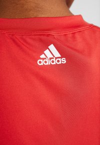 Červené sportovní tričko s hladkým povrchem, kulatým výstřihem a bílým logem Adidas na zadní straně. Materiál vypadá lehce a prodyšně.