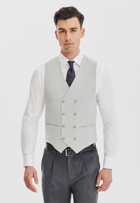 Jasnoszara, teksturowana kamizelka w kształcie litery V, z podwójnymi guzikami i dopasowanym krojem, noszona na białej koszuli i szarych spodniach.