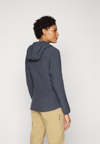 Mørkegrå hættejakke med struktureret bagpanel, tætsiddende design, lange ærmer og blødt stof. Kombineret med lyse khaki-bukser.