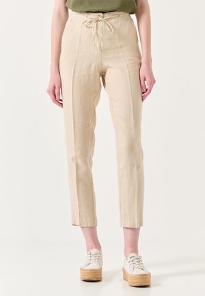 Beige Leinenhosen mit gelockertem Schnitt, verstellbarem Taillenbund und schmal zulaufenden Beinen. Kombiniert mit weißen Plateausneakern für einen Kontrast.