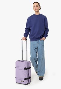 Fialový kufr na kolečkách se dvěma černými popruhy, teleskopickou rukojetí a hladkým povrchem. Model nosí modrou mikinu a světlé džíny.