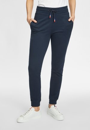 Persoon draagt marineblauwe joggingbroek met trekkoord en witte sneakers met rode, witte en blauwe streepdetails, handen in de zakken.