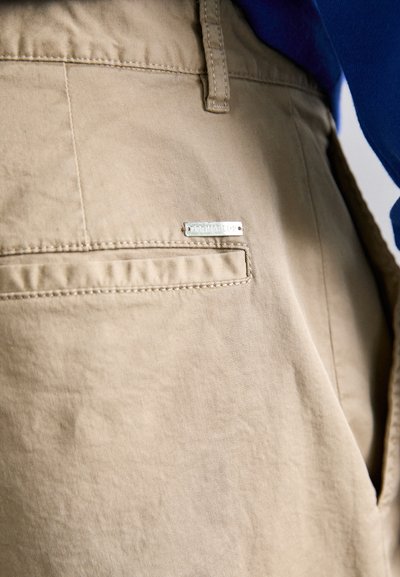 Pantaloni beige realizzati in tessuto liscio, dotati di un piccolo tag metallico con logo. Inclusi tasche laterali con dettagli di cucitura ordinata.
