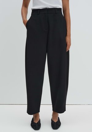 Persoon draagt hoge, losse zwarte broek gecombineerd met zwarte platte schoenen met plooien en een wit topje in de broek gestopt, staat met handen in de zakken.