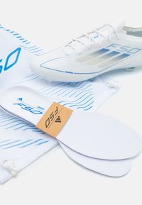Boksa s bijelim nogometnim kopačkama s plavim akcentima i teksturiranim gornjim dijelom, uz bijele uloške i vrećicu za nošenje sa plavim valovitim uzorcima.