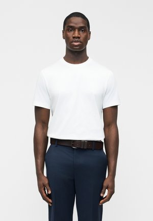 Moški stoji obrnjeno naprej, oblečen v preprosto belo srajco s kratkimi rokavi, temno modre hlače in temno rjav pas, na enobarvnem ozadju.