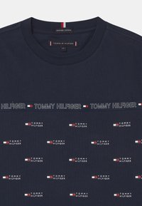 Navy blue organic cotton t-shirt featuring the brand name "TOMMY HILFIGER" in repeated white text and small logo accents. Crew neckline.