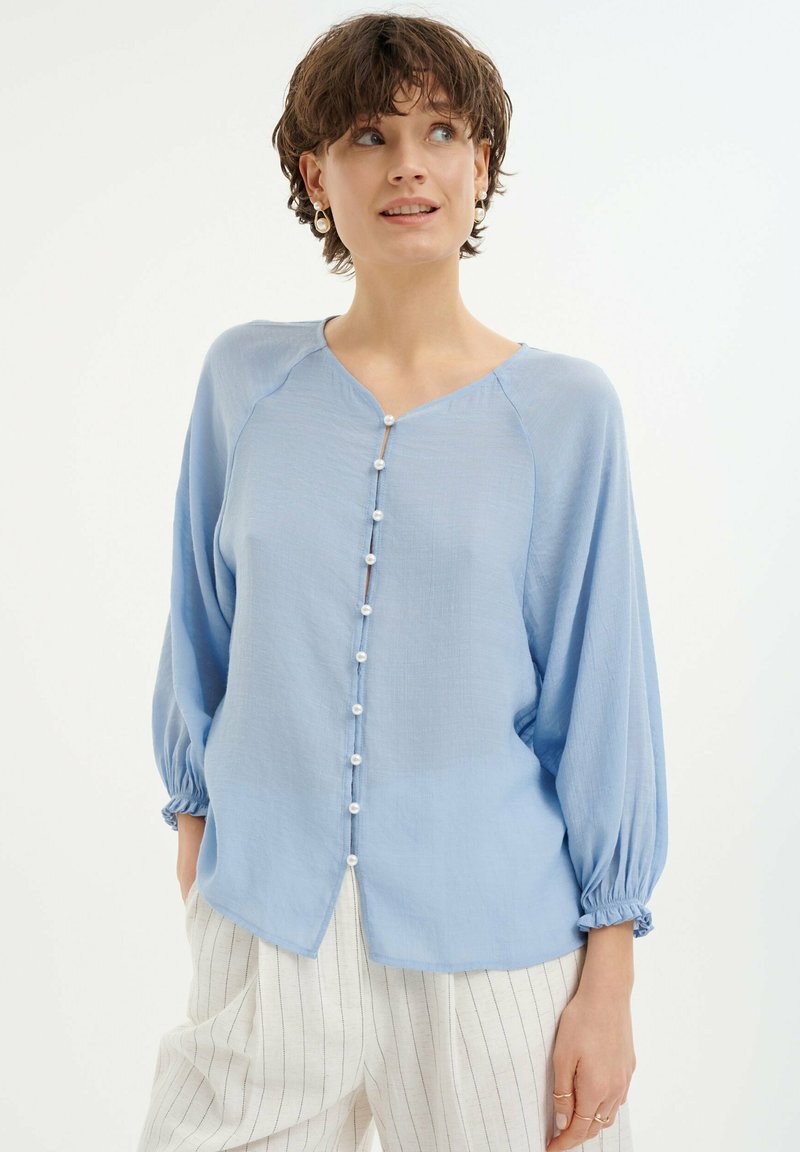 Hellbluse mit Knopfleiste, Raglanärmeln und elastischen Manschetten. Weiches, strukturiertes Material mit kleinen weißen Knöpfen entlang der Front.