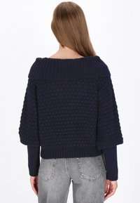 Maglione lavorato a maglia blu navy con un design a punto bolla texturizzato, ampio colletto a coste e maniche corte, abbinato a pantaloni grigi.
