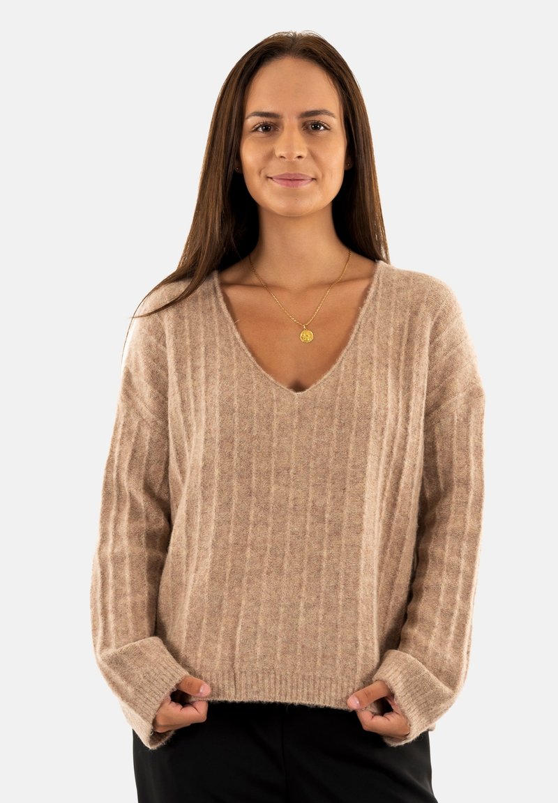 Pull en tricot beige avec un design en V, texture côtelée, épaules tombantes et coupe décontractée. Présente des manches longues et une apparence douce.