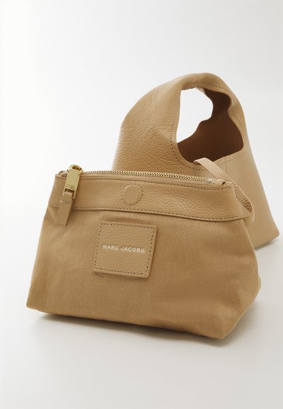 Beige laukku, jossa on teksturoitu materiaali, pyöreä kahva, etutasku vetoketjulla ja labeli, jossa on painettu valkoisella "MARC JACOBS".