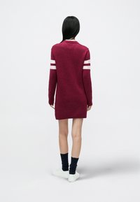 Bordó kötött pulóverruha fehér csíkokkal az ujjain, kerek nyakkivágással, combközépig érő hosszúsággal és bordázott szegéllyel; a modell sötét zoknit és fehér tornacipőt visel.