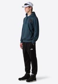 Donkerblauwe sweatshirt met een capuchon en een voorkantzak, gecombineerd met zwarte joggingsbroek met een wit logo, en zwarte wandelschoenen.