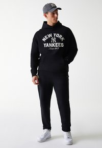 Sweatshirt preto com o gráfico "NEW YORK YANKEES", calças de fato pretas a condizer e ténis brancos. Um boné cinzento com padrão completa o visual.