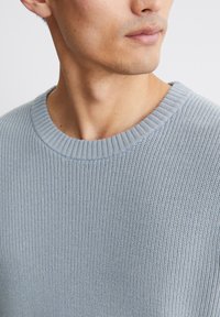Hellblauer Strickpullover mit geripptem Rundhalsausschnitt und strukturiertem Stoff, mit einem Nahaufnahme-Fokus auf dem Nacken- und Schulterbereich.