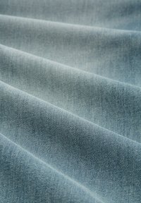 Jasnoniebieska tkanina dżinsowa o gładkiej fakturze, pokazująca diagonalne sploty skośne i miękkie fałdy. Powierzchnia ma delikatny połysk.